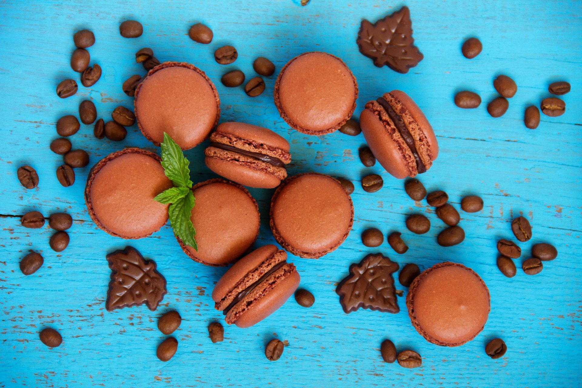Close-up of chocolate macarons and coffee beans scattered on a bright blue wooden surface, with pieces of chocolate and a sprig of mint, in vibrant 4K Ultra HD detail.