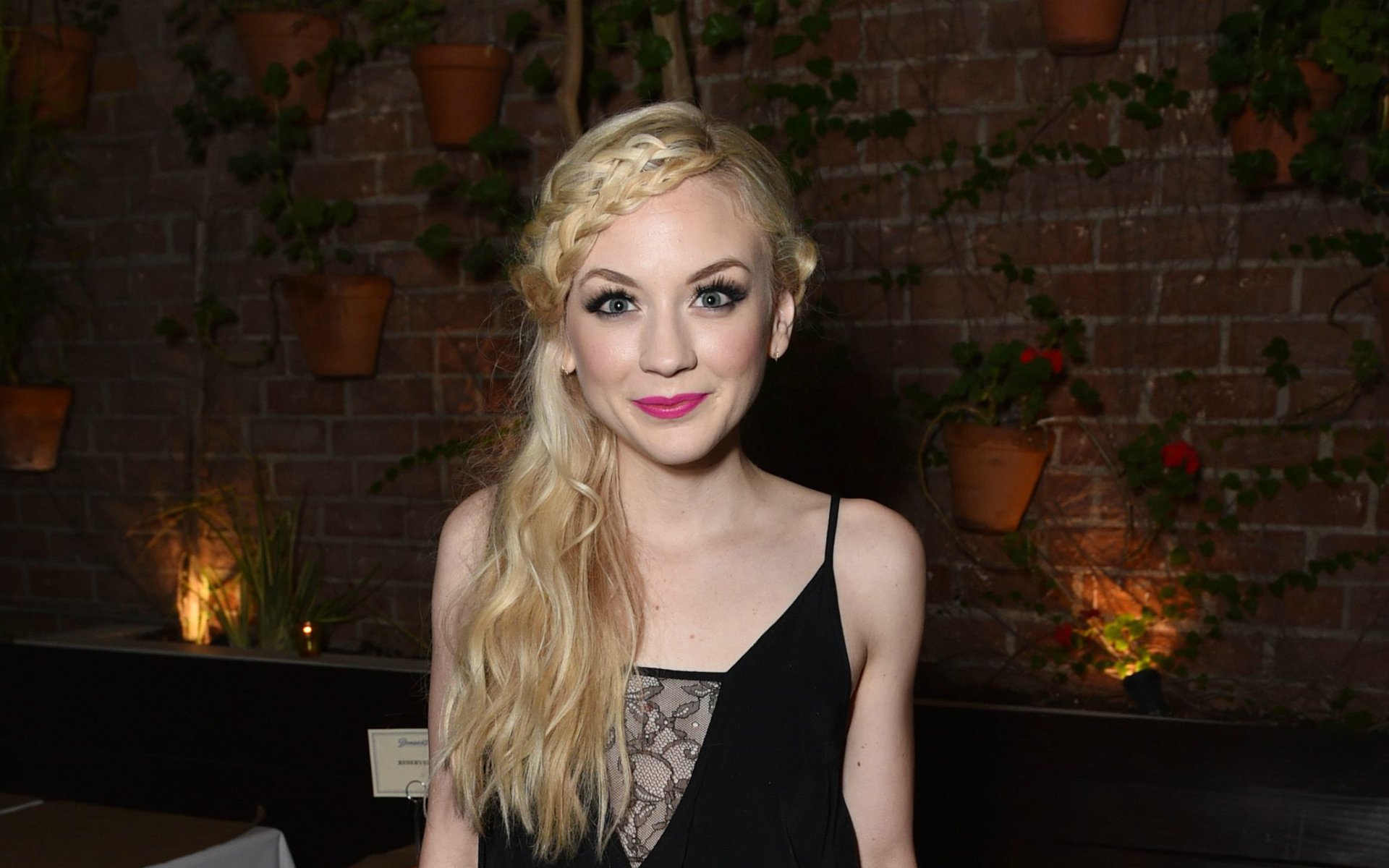 HD desktop wallpaper featuring Emily Kinney, American singer and actress, against a softly lit brick wall with potted plants in the background.