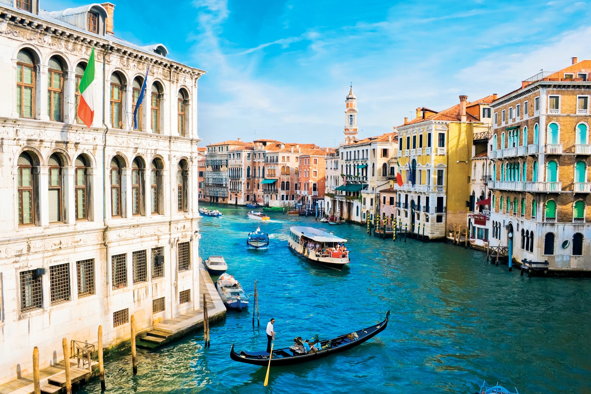 4K Ultra HD view of Venice's iconic canal with gondolas and colorful historic buildings under a bright blue sky, showcasing the charm of this man-made Italian cityscape.