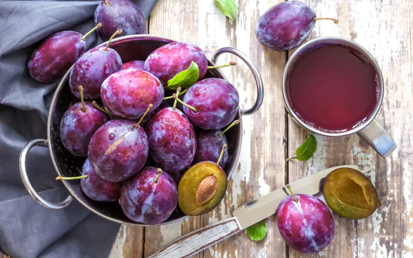 A vibrant 4K Ultra HD desktop wallpaper featuring fresh purple plums in a bowl beside a cup of plum juice on a rustic wooden surface.