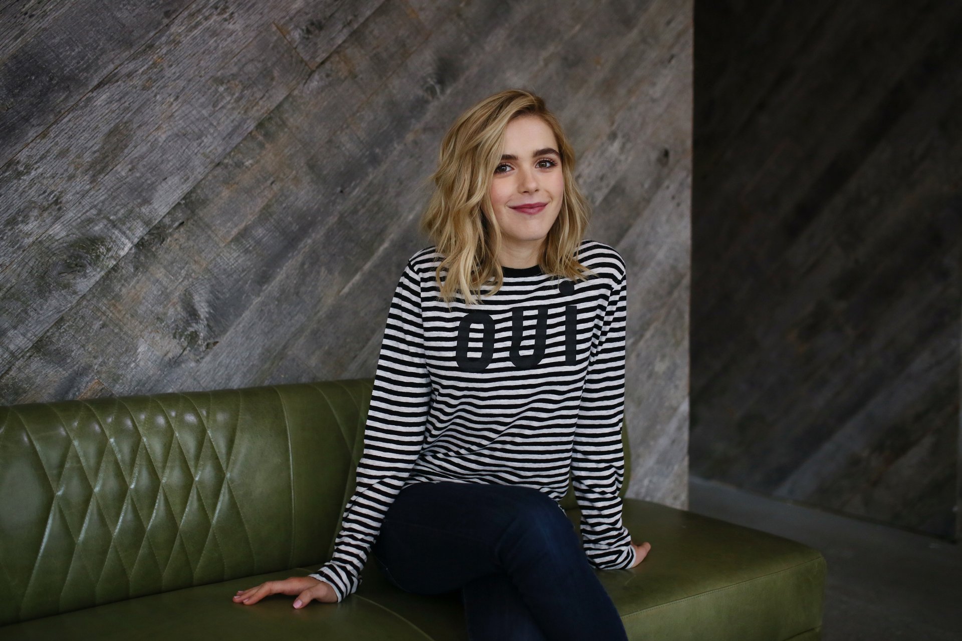 HD PC desktop wallpaper of celebrity Kiernan Shipka sitting on a green leather bench against a textured gray wall, wearing a black-and-white striped sweater.