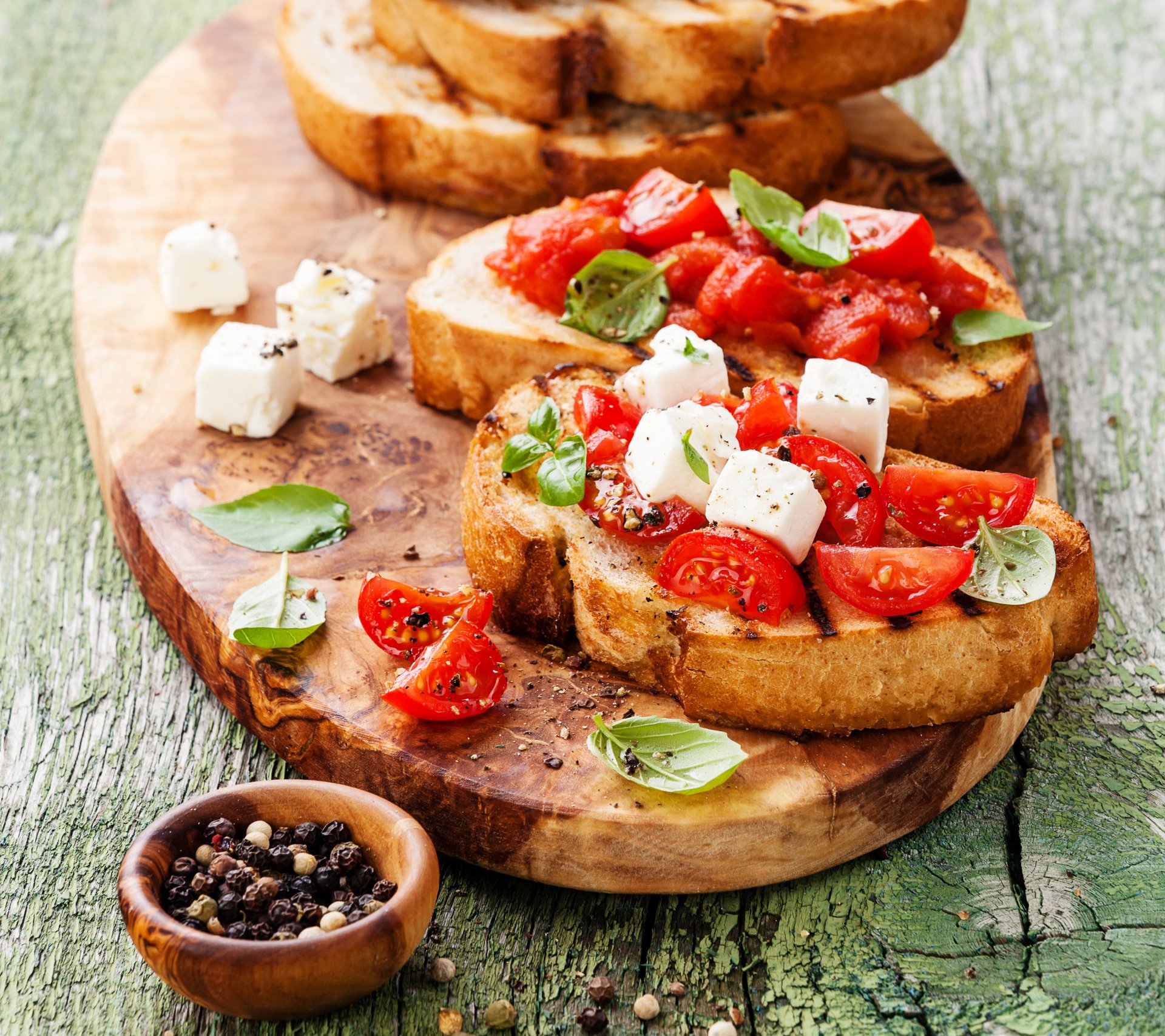 Close-up of a sandwich with fresh tomato, cheese cubes, basil leaves, and cracked pepper on rustic bread, set on a wooden board with a peppercorn bowl.