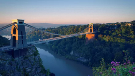 Scenic HD desktop wallpaper featuring the Clifton Suspension Bridge spanning the Avon Gorge in Bristol, surrounded by lush greenery and a calm river at sunrise.