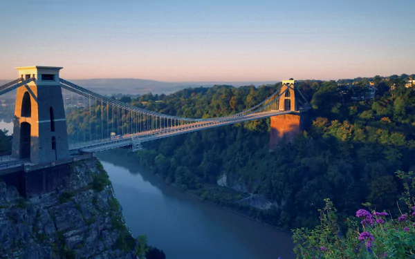Scenic HD desktop wallpaper featuring the Clifton Suspension Bridge spanning the Avon Gorge in Bristol, surrounded by lush greenery and a calm river at sunrise.