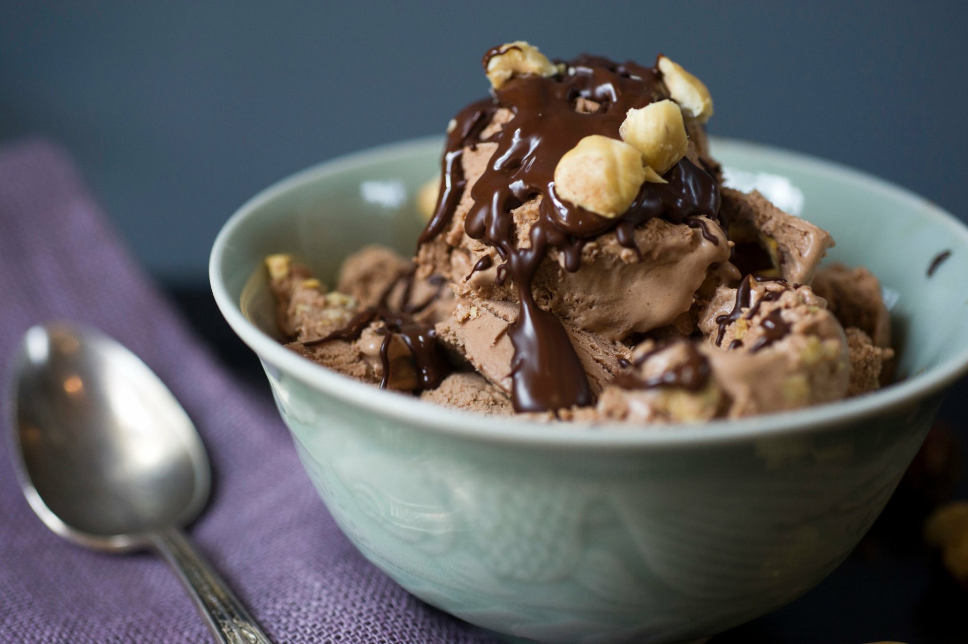 A bowl of creamy chocolate ice cream topped with chocolate sauce and nuts, placed on a purple cloth with a spoon beside it. HD desktop wallpaper and background.