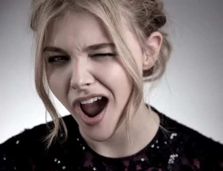 HD desktop wallpaper featuring Celebrity Chloë Grace Moretz winking with an expressive look on her face, set against a neutral background.