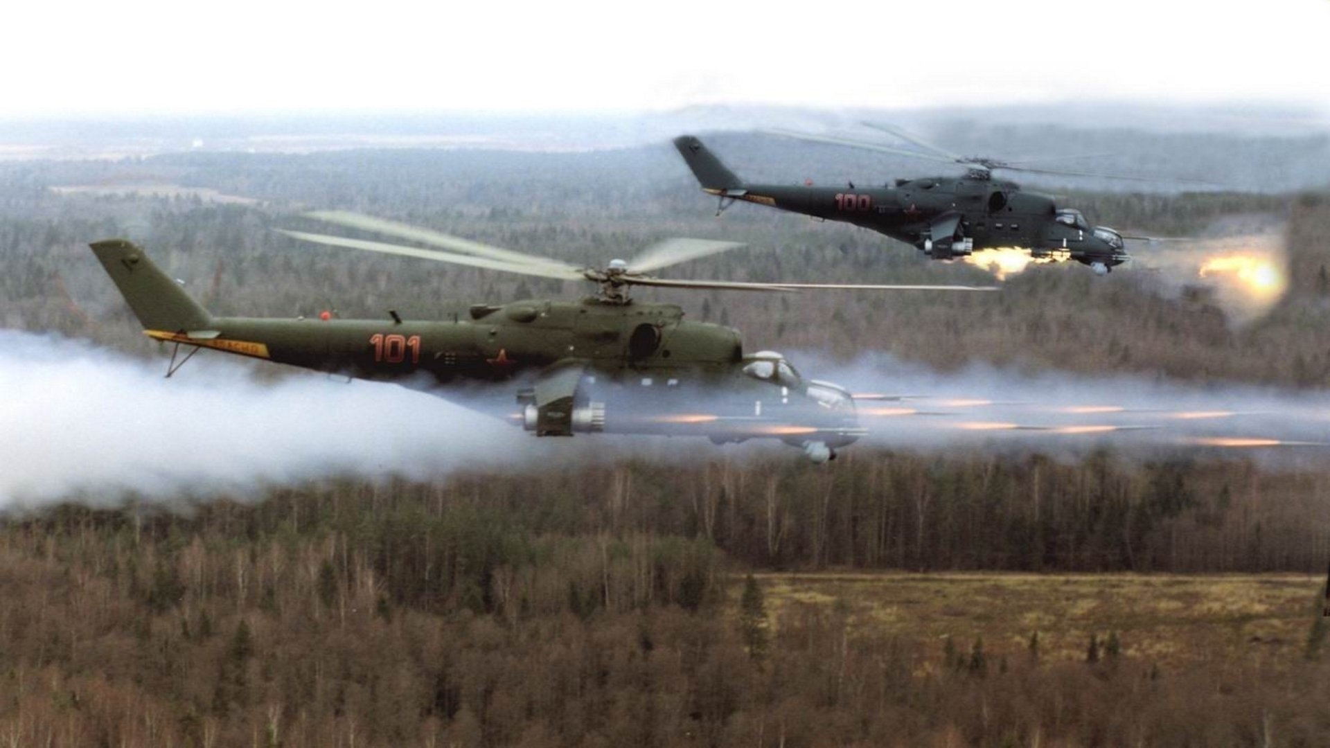 HD desktop wallpaper featuring two military Mil Mi-24 helicopters flying low over a forested landscape with visible rotor motion and environmental haze.