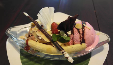 HD desktop wallpaper of a banana split dessert featuring scoops of ice cream, chocolate curls, nuts, strawberry, and whipped cream on a dark wooden table.