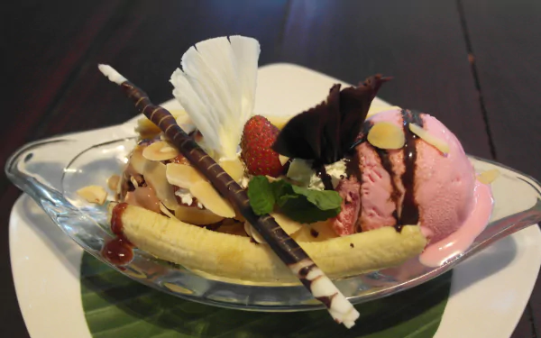 HD desktop wallpaper of a banana split dessert featuring scoops of ice cream, chocolate curls, nuts, strawberry, and whipped cream on a dark wooden table.