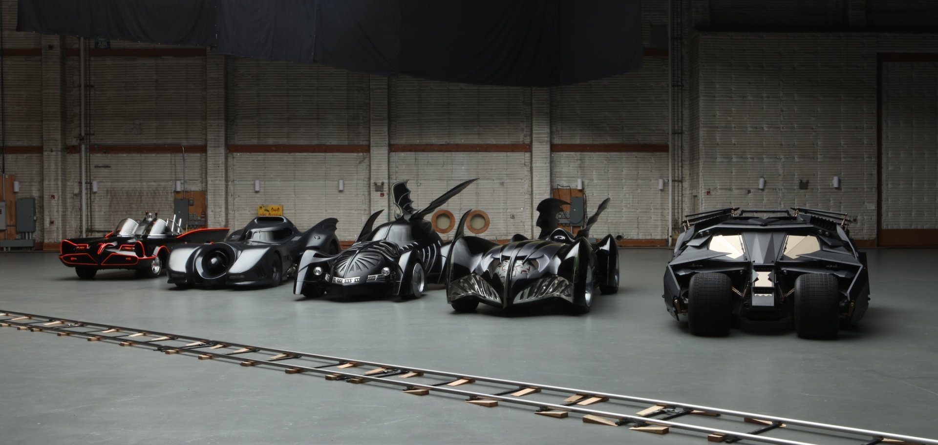 HD PC desktop wallpaper featuring a lineup of Batmobile vehicles in a spacious industrial garage setting.