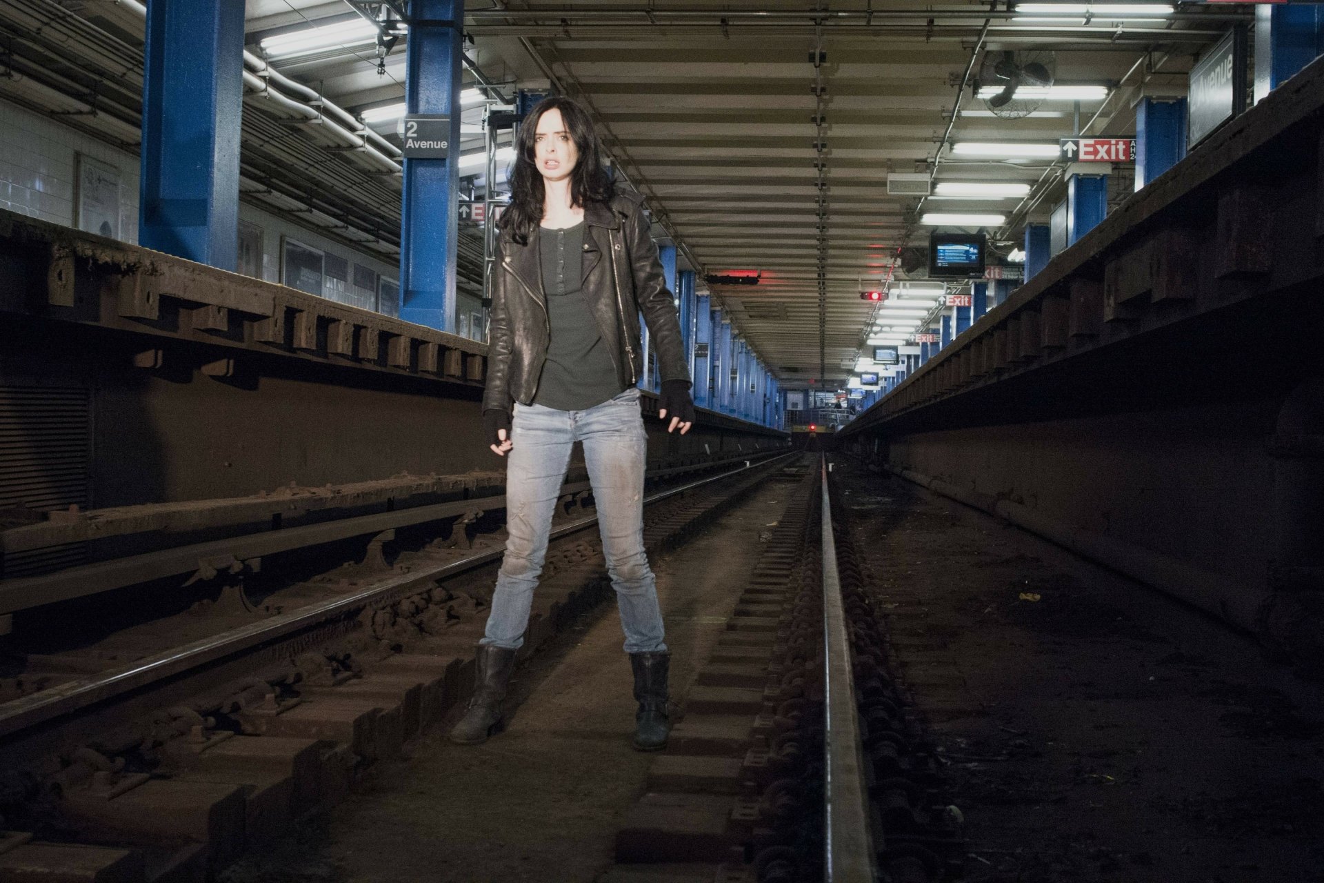 Krysten Ritter as Jessica Jones stands confidently on subway tracks in a dimly lit tunnel, captured in a 4K Ultra HD TV show wallpaper background.