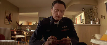 HD PC desktop wallpaper: movie still of an actor in military dress uniform seated at a kitchen table, solemnly handling a small object in a warmly lit living-room setting.