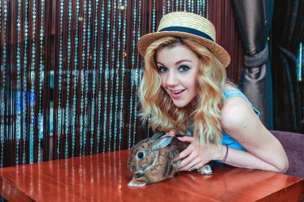 Blonde Russian singer Yulianna Karaulova with brown eyes smiles while holding a rabbit, wearing a straw hat against a shimmering curtain backdrop.