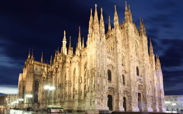 Night view of Milan Cathedral in Italy, illuminated against a deep blue sky, showcasing its intricate religious Gothic architecture in stunning 4K Ultra HD detail.
