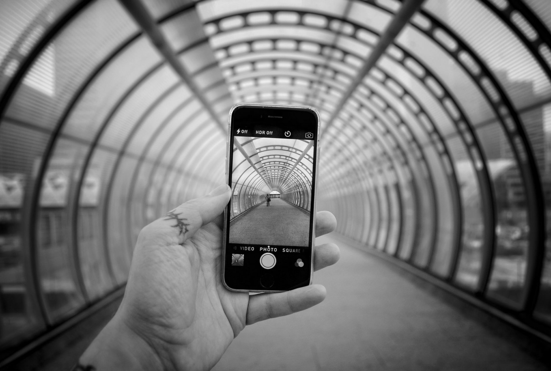 Black and white HD desktop wallpaper showing a hand holding an Apple iPhone, capturing a tunnel with a geometric, repetitive arch design.