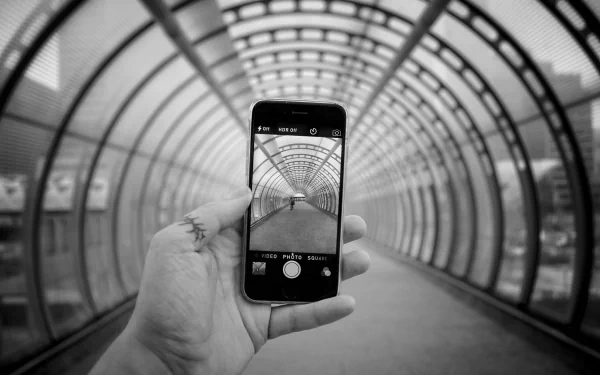 Black and white HD desktop wallpaper showing a hand holding an Apple iPhone, capturing a tunnel with a geometric, repetitive arch design.