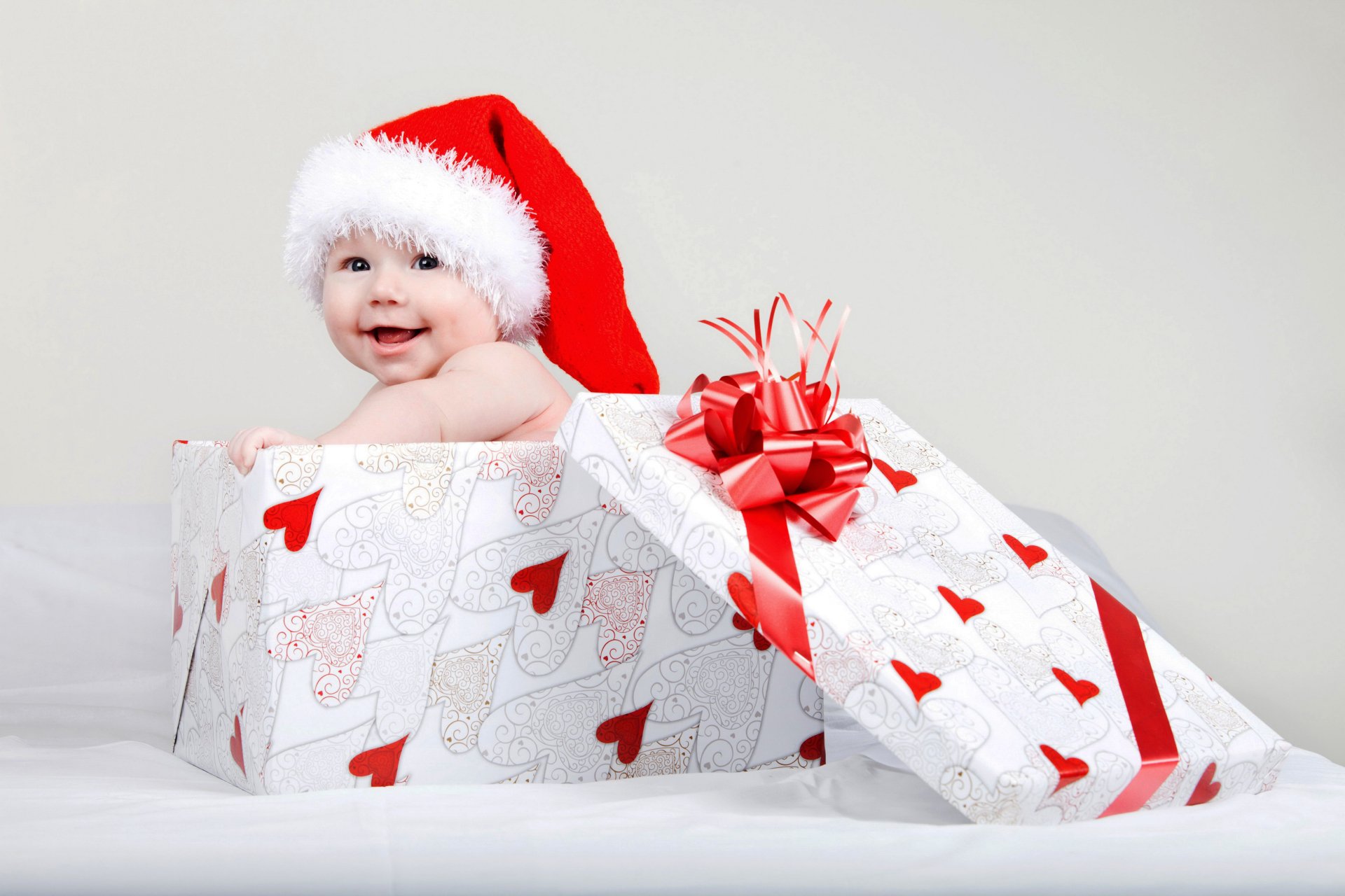 A baby wearing a Santa hat smiles while sitting inside a large Christmas gift box wrapped in white paper with red hearts and ribbons, captured in 4K Ultra HD.