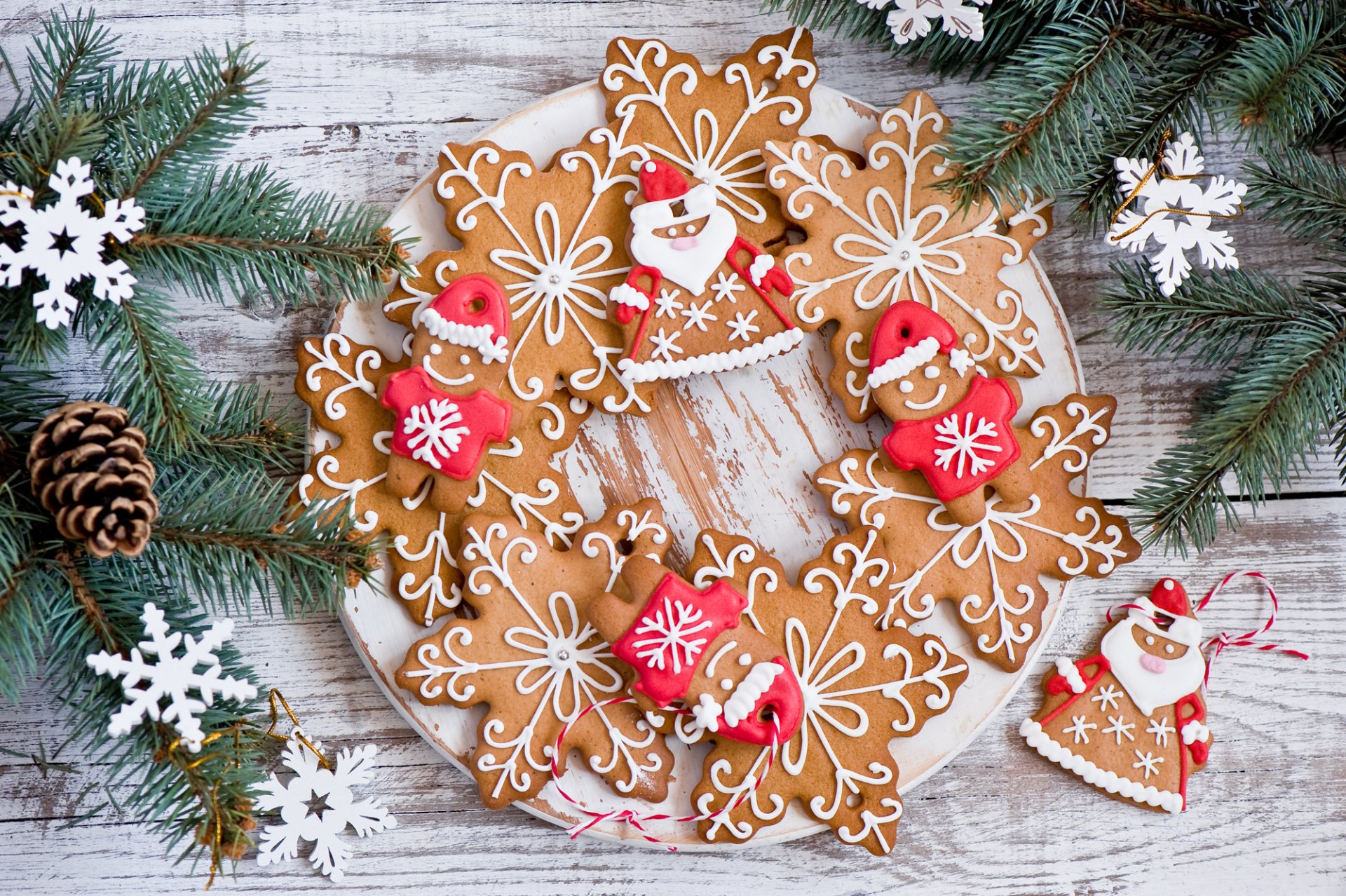 Holiday-themed gingerbread cookies shaped like snowflakes, reindeer, and Santa Claus arranged in a wreath on a rustic wooden background with pine branches and snowflake decorations.