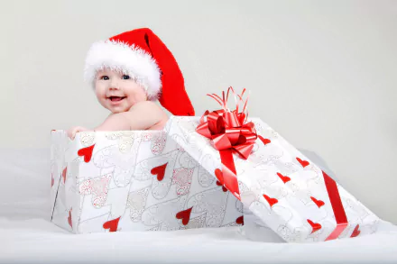 A baby wearing a Santa hat smiles while sitting inside a large Christmas gift box wrapped in white paper with red hearts and ribbons, captured in 4K Ultra HD.