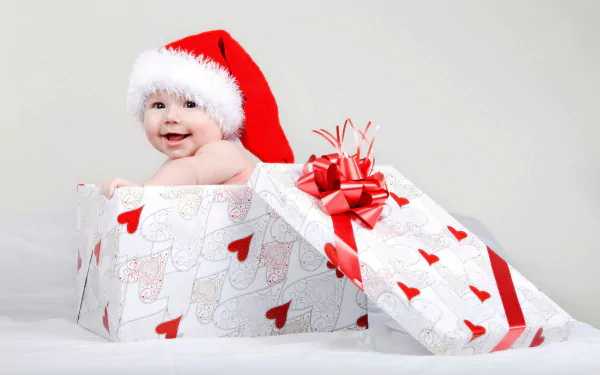 A baby wearing a Santa hat smiles while sitting inside a large Christmas gift box wrapped in white paper with red hearts and ribbons, captured in 4K Ultra HD.
