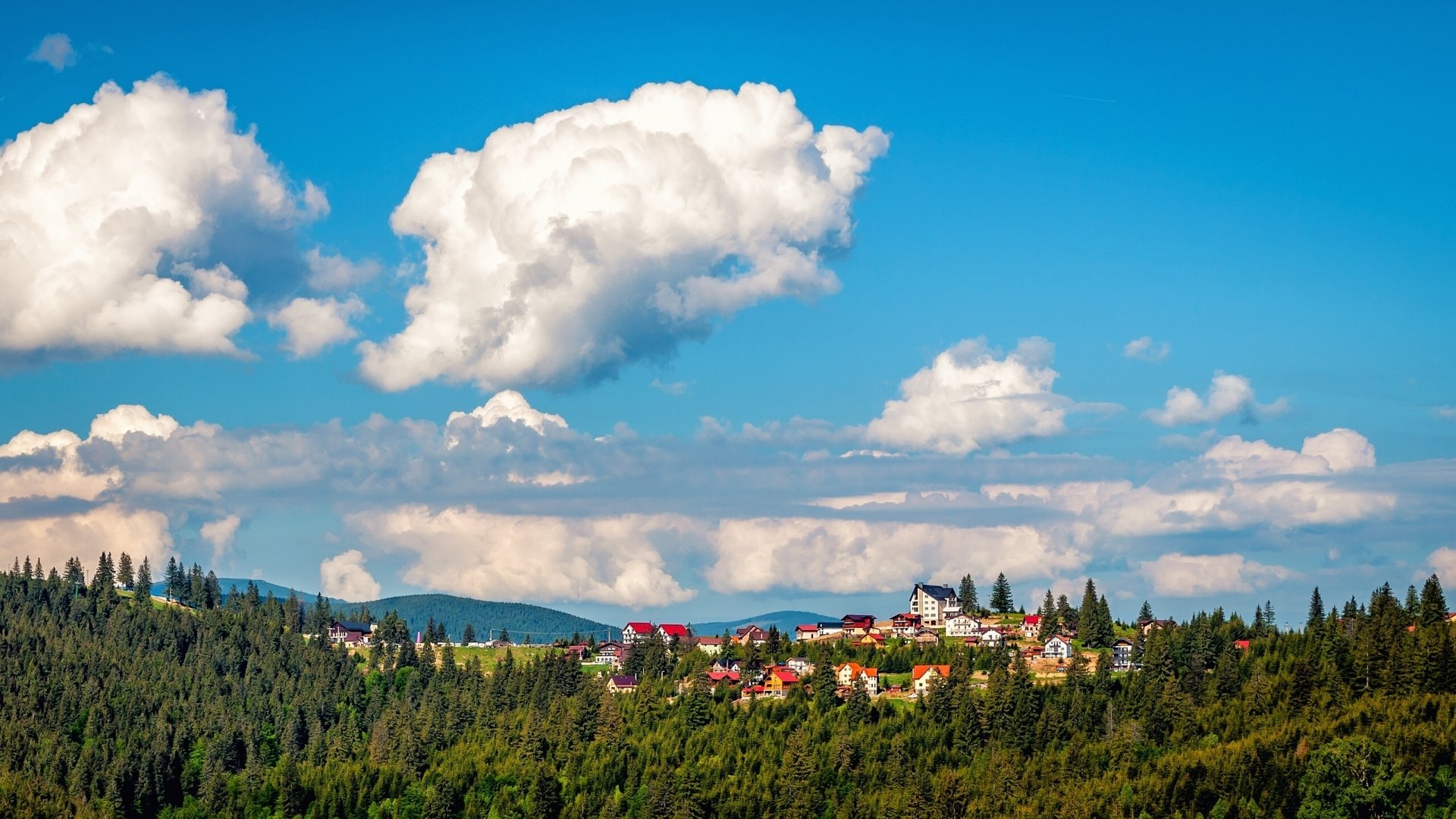 HD desktop wallpaper showing a Romanian village nestled among lush green hills under a vibrant blue sky with large, fluffy white clouds.