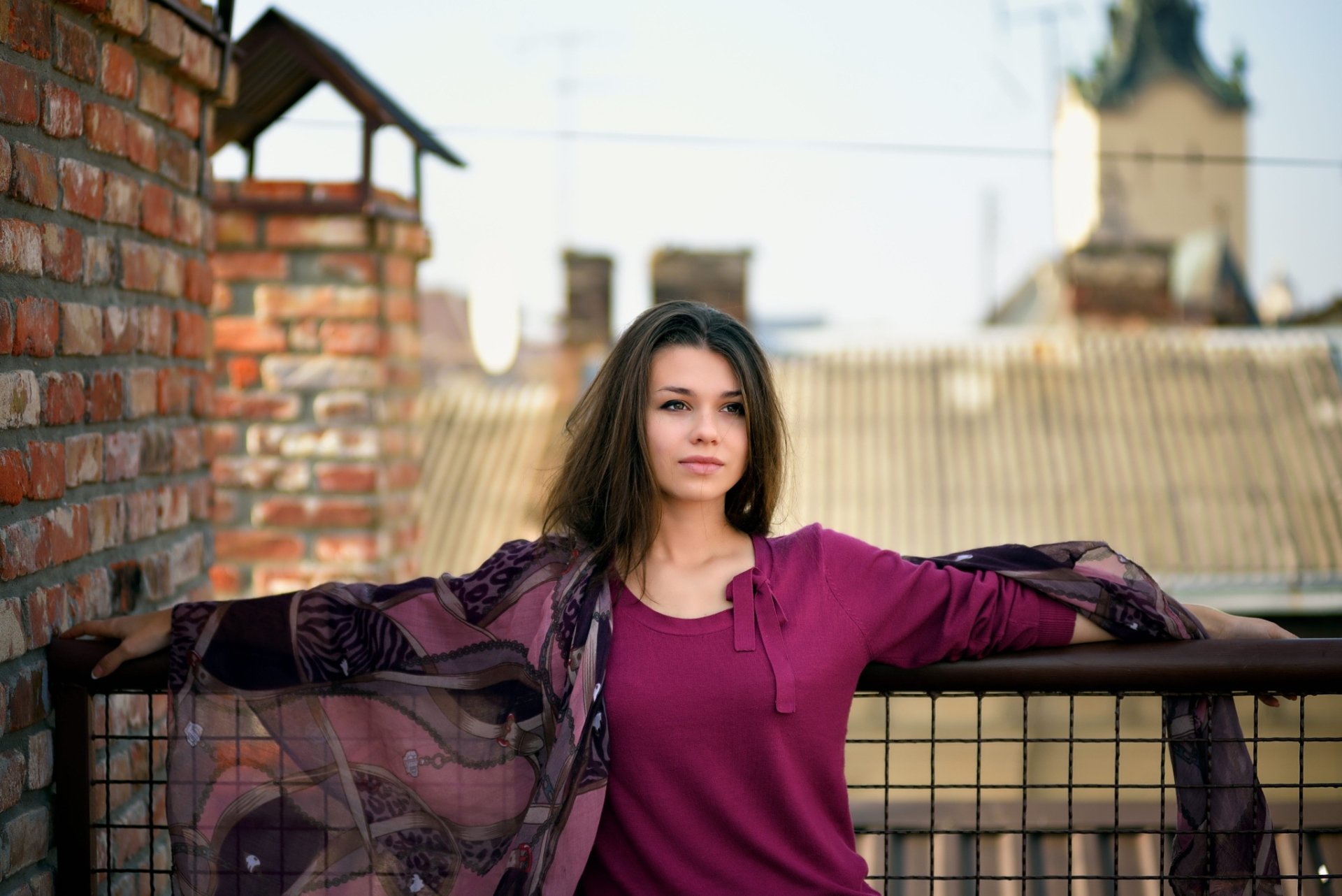 A woman in a purple top stands confidently on a rooftop terrace with her arms outstretched, framed by rustic brick walls. HD desktop wallpaper and background with a model.