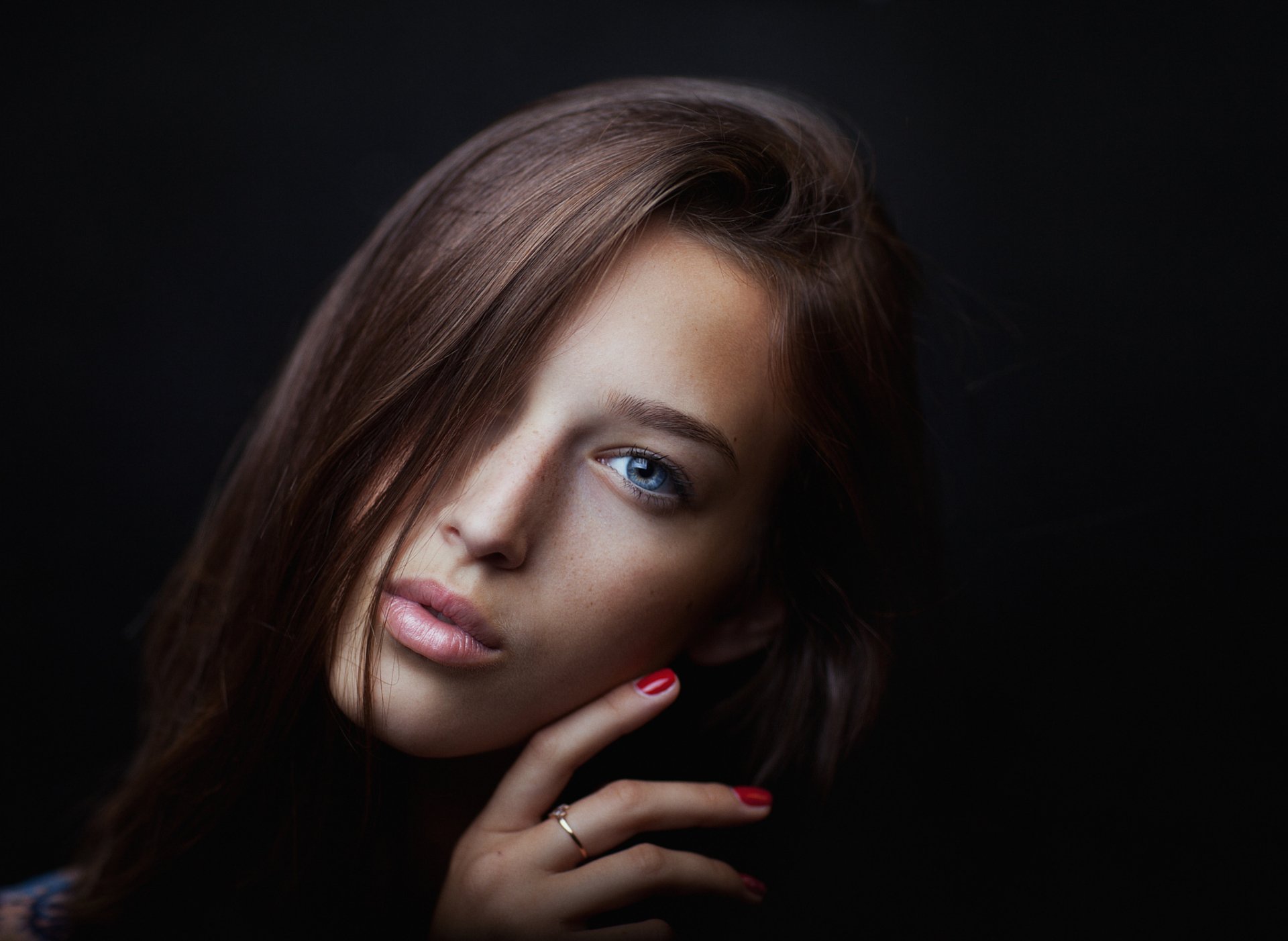 A brunette model with long hair and striking blue eyes gazes calmly into the camera, her hand poised thoughtfully near her face against a dark background.