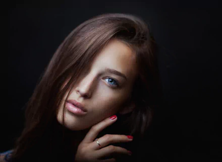 A brunette model with long hair and striking blue eyes gazes calmly into the camera, her hand poised thoughtfully near her face against a dark background.