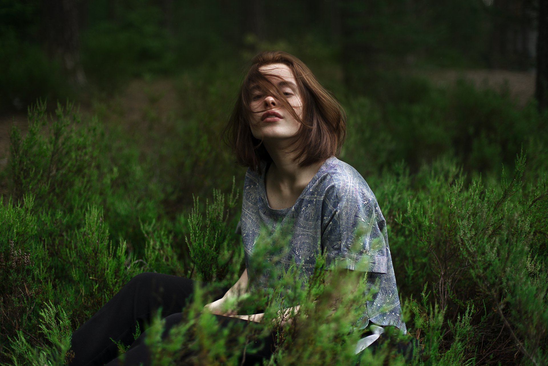 A brunette woman outdoor model sits among green foliage with soft bokeh in the background, captured in a moody HD desktop wallpaper and background.