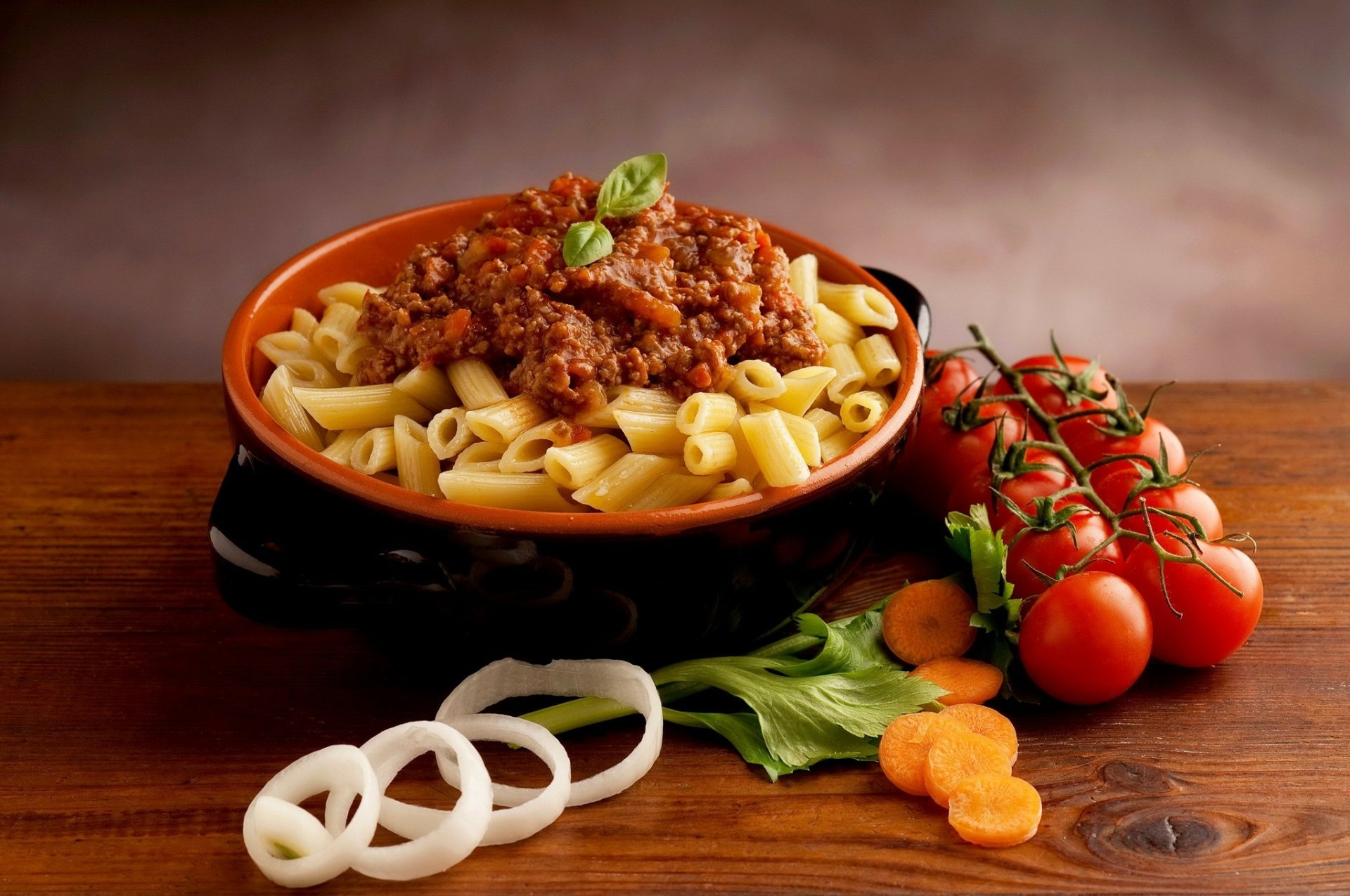 HD desktop wallpaper featuring a bowl of pasta topped with tomato meat sauce, surrounded by fresh tomatoes, basil, onion rings, and carrot slices on a wooden surface.
