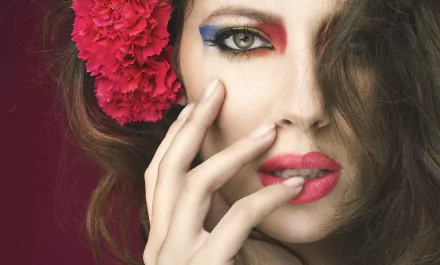Close-up HD wallpaper of a brunette woman with green eyes, bold red lipstick, colorful eye makeup, a red flower in her hair, and her hand near her face.