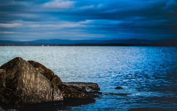 5K Ultra HD PC Desktop Wallpaper and Background: nature rock on a serene blue shoreline, rippling water and dramatic cloud-streaked sky meeting the distant horizon.