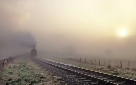 A steam train emerges through a thick fog on a tranquil railroad, with a soft sunrise illuminating the scene, creating a captivating HD desktop wallpaper.