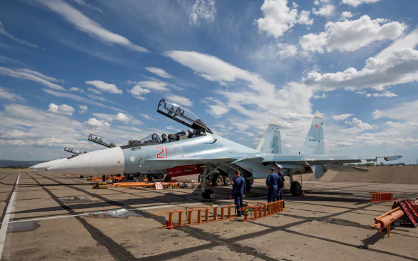 HD desktop wallpaper featuring the military aircraft Sukhoi Su-30. The plane is stationed on a tarmac under a partly cloudy sky, with maintenance crew members visible around it.