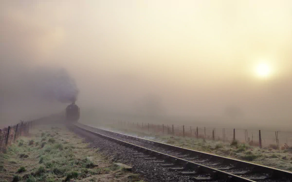 A steam train emerges through a thick fog on a tranquil railroad, with a soft sunrise illuminating the scene, creating a captivating HD desktop wallpaper.