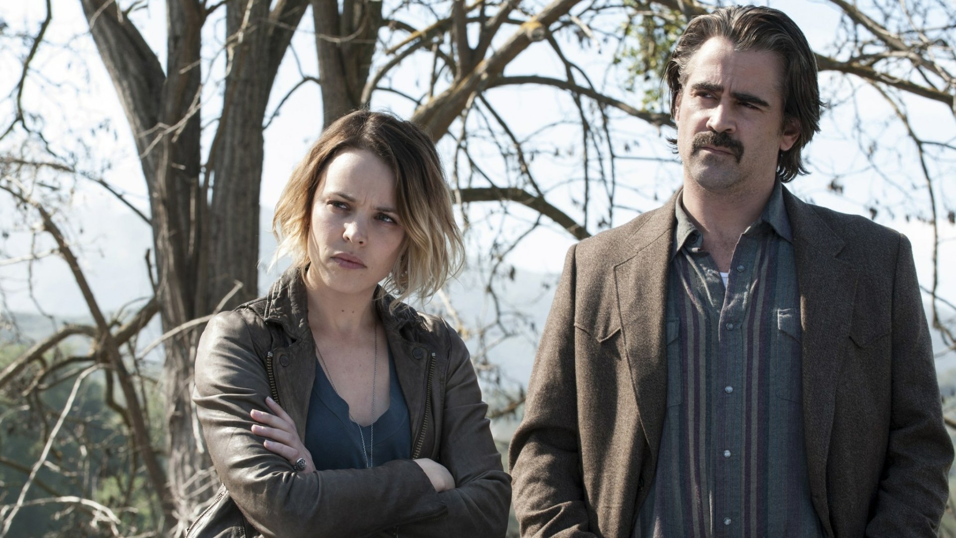 HD desktop wallpaper: two actors as detectives in the TV show True Detective — a woman with arms folded and a mustached man standing in front of bare trees.