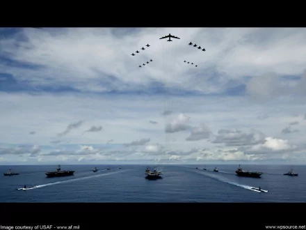 A stunning HD wallpaper showcasing a military aircraft carrier amidst the ocean, with formations of jets flying above and naval vessels navigating the water, set against an expansive sky.