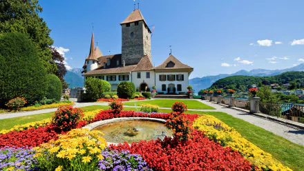 A vibrant garden with colorful flowers and a fountain in front of a historic castle building in Switzerland, set against a clear blue sky and mountain backdrop.