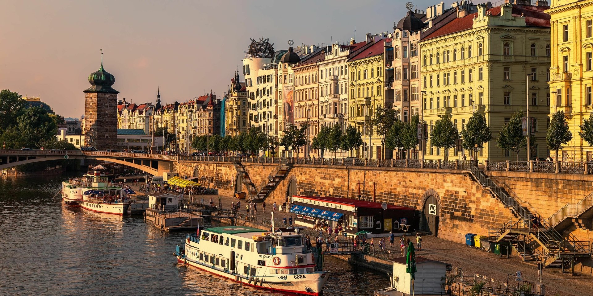 Man-made Prague riverside with historic facades, boats along the Vltava and a sunlit quayside — 2K Quad HD PC desktop wallpaper and background.