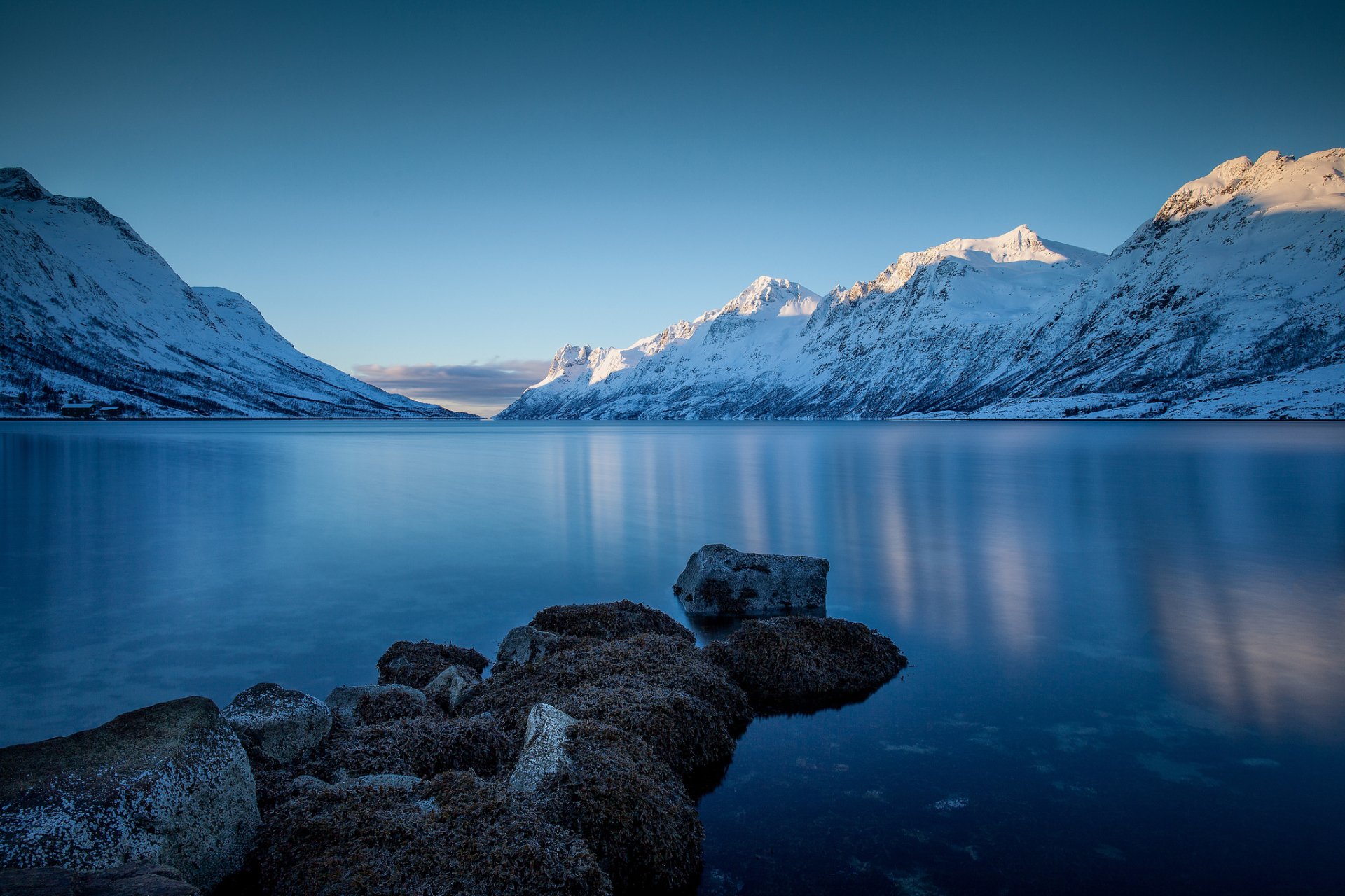 A serene winter landscape with snow-covered mountains reflecting in a tranquil lake, surrounded by rocky shores, creating a stunning HD desktop wallpaper for nature lovers.