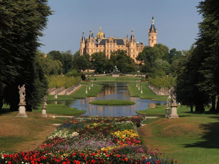 Schwerin Palace in Germany stands behind a formal garden with colorful flowers, statues, and a reflecting pool under a clear sky in this HD desktop wallpaper.