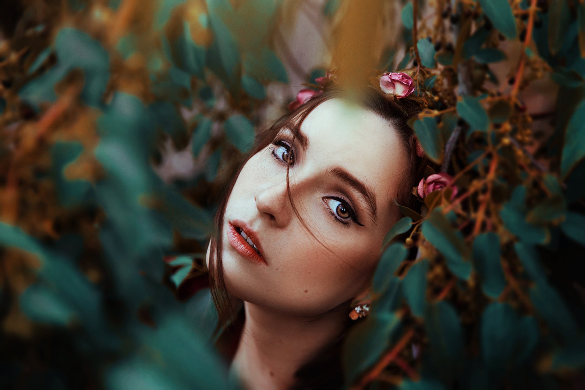 HD desktop wallpaper featuring a brunette woman with brown eyes, wearing lipstick and floral earrings, framed by foliage with a soft bokeh background.