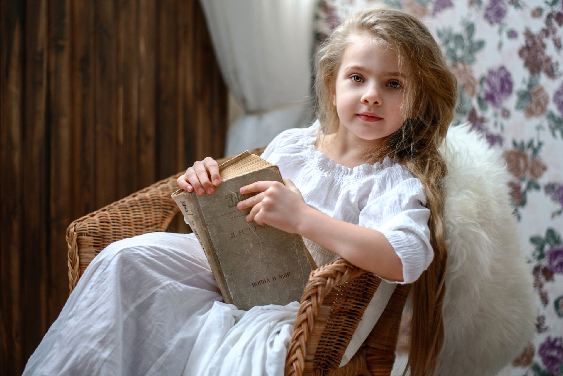 A little girl with green eyes and blonde hair sits in a wicker chair, holding an old book. The cozy setting features floral wallpaper, capturing a tranquil moment in photography.