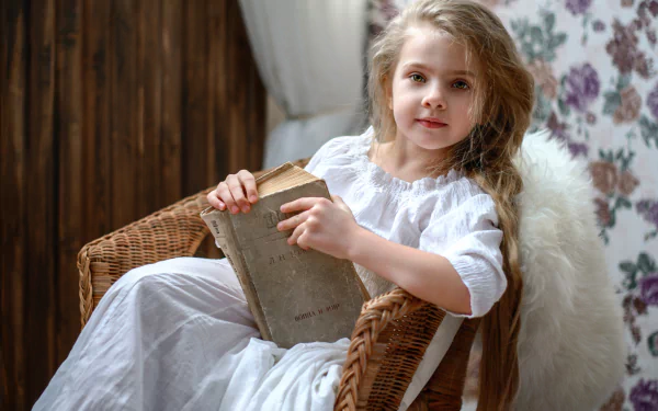 A little girl with green eyes and blonde hair sits in a wicker chair, holding an old book. The cozy setting features floral wallpaper, capturing a tranquil moment in photography.