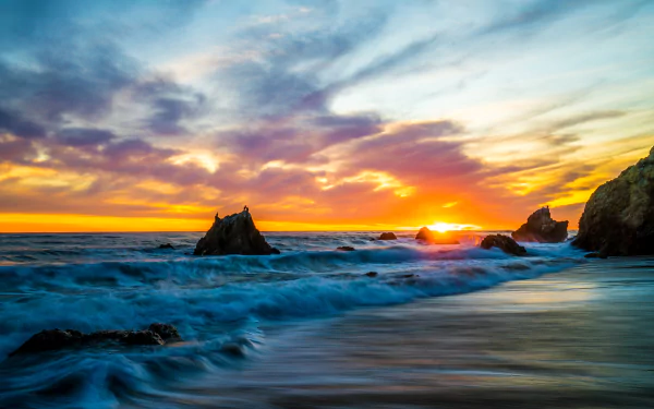 Vibrant 8K Ultra HD wallpaper of a sunset over the ocean, showcasing waves crashing against rocks on a beach under a colorful sky.