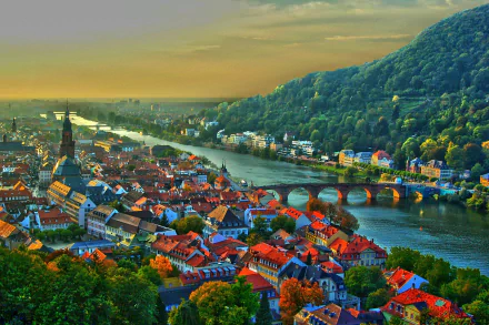 A vibrant cityscape of Heidelberg, Germany, featuring historic buildings, a stone bridge over the river, and lush green hills under a golden sunset sky.