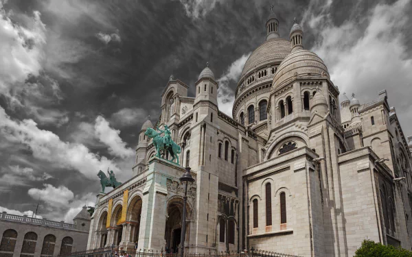 HD desktop wallpaper featuring the Sacré-Cœur Basilica in Paris, France, with dramatic clouds and a prominent religious statue under a dynamic sky.