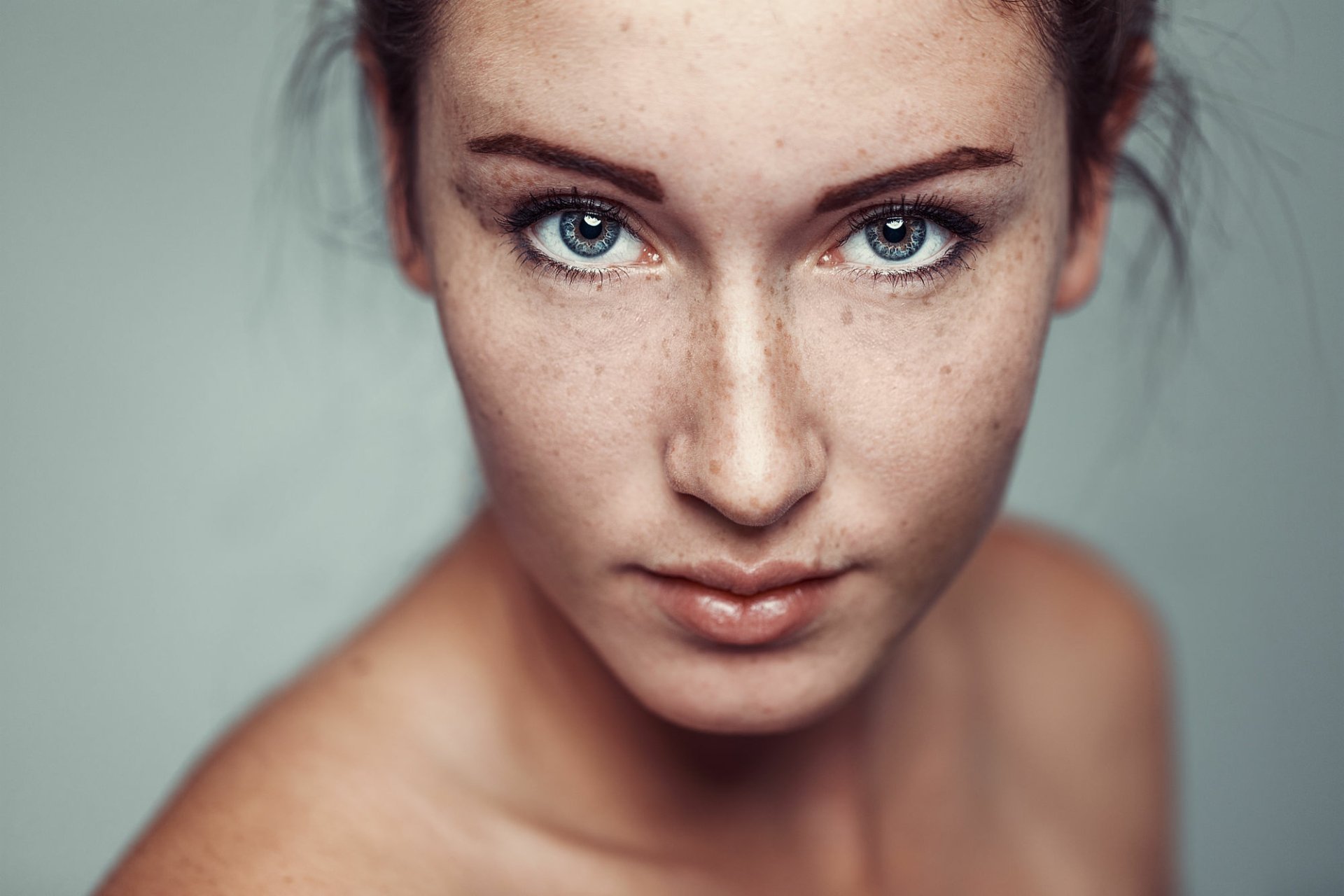Close-up HD portrait of a female model with blue eyes, visible freckles, and a focused expression, set against a soft, neutral background.