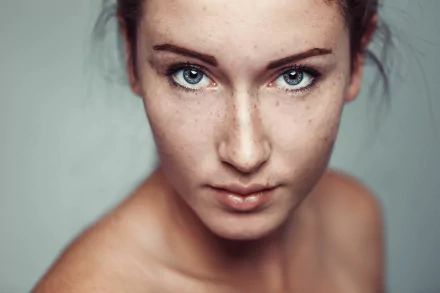 Close-up HD portrait of a female model with blue eyes, visible freckles, and a focused expression, set against a soft, neutral background.