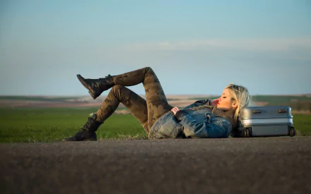 Blonde woman lying on a road beside a silver suitcase, contemplative mood; 2K Quad HD PC desktop wallpaper background.